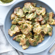 A blue plate of smashed potatoes topped with vibrant green pesto sits on a white surface next to a bowl of sauce with a small whisk and a beige cloth napkin.