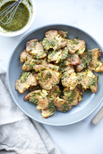 A blue plate of smashed potatoes topped with vibrant green pesto sits on a white surface next to a bowl of sauce with a small whisk and a beige cloth napkin.
