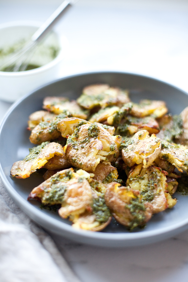 A gray plate with smashed potatoes, perfectly roasted and topped with vibrant green herb sauce. In the background, a white bowl containing more green sauce and a spoon completes the scene.