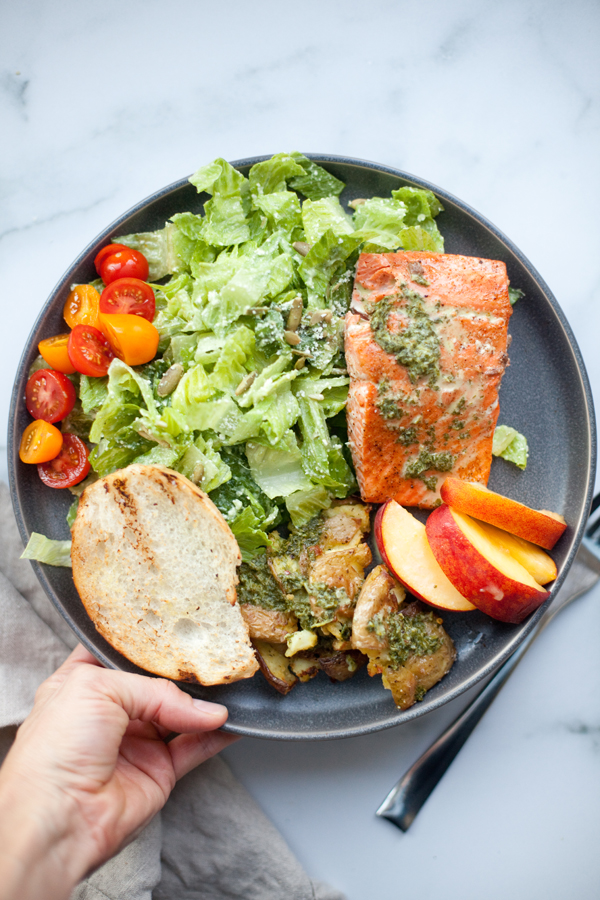 A plate with grilled salmon topped with green sauce, a green salad with cherry tomatoes, smashed potatoes, peach slices, and a slice of toasted bread. A hand holds the plate on a marble surface.