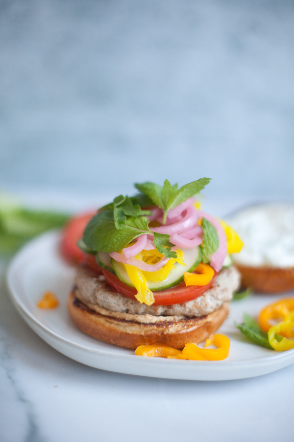 Open-faced Greek Turkey Burger on a bun with tomato, cucumber, yellow bell pepper, pickled onions, and fresh mint, served on a white plate with a side of creamy sauce.