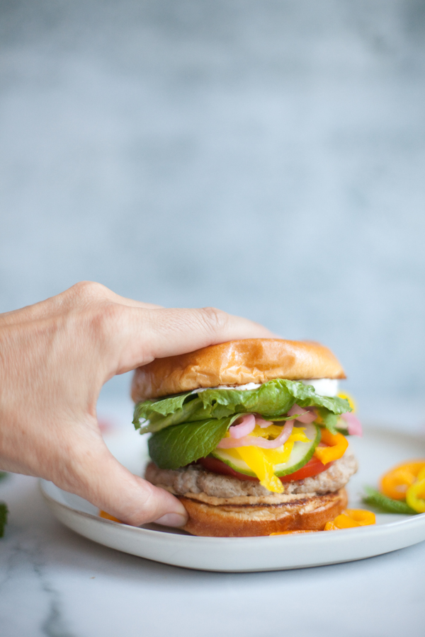 A hand reaches to pick up a Greek Turkey Burger with lettuce, tomato, pickles, onions, and sauce on a brioche bun, placed on a white plate with yellow pepper slices nearby.