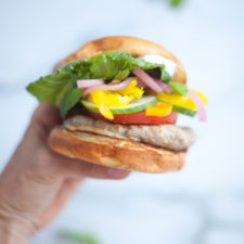 A hand holds a Greek Turkey Burger sandwich with lettuce, tomato, cucumber, pickled onions, yellow peppers, and a patty in a bun, against a blurred light background.