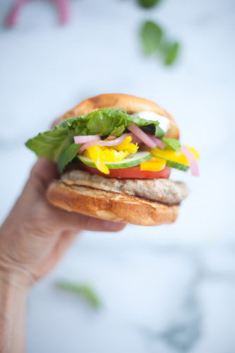 A hand holds a Greek Turkey Burger sandwich with lettuce, tomato, cucumber, pickled onions, yellow peppers, and a patty in a bun, against a blurred light background.
