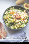 A bowl of chopped salad with lettuce, avocado, corn, cheese, salsa, and dressing sits on a table next to a wooden spoon and a blue napkin. Text reads: Copycat Factory Chopped Salad—your homemade take on the beloved Skinnylicious favorite.