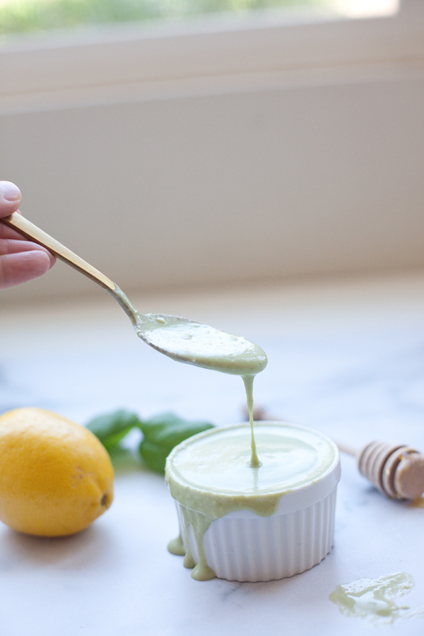 A hand holds a spoon with green sauce dripping into a small white ramekin, evoking the fresh flavors of a Cobb salad with creamy lemon basil dressing. A lemon, basil leaves, and a honey dipper are nearby on a white surface.