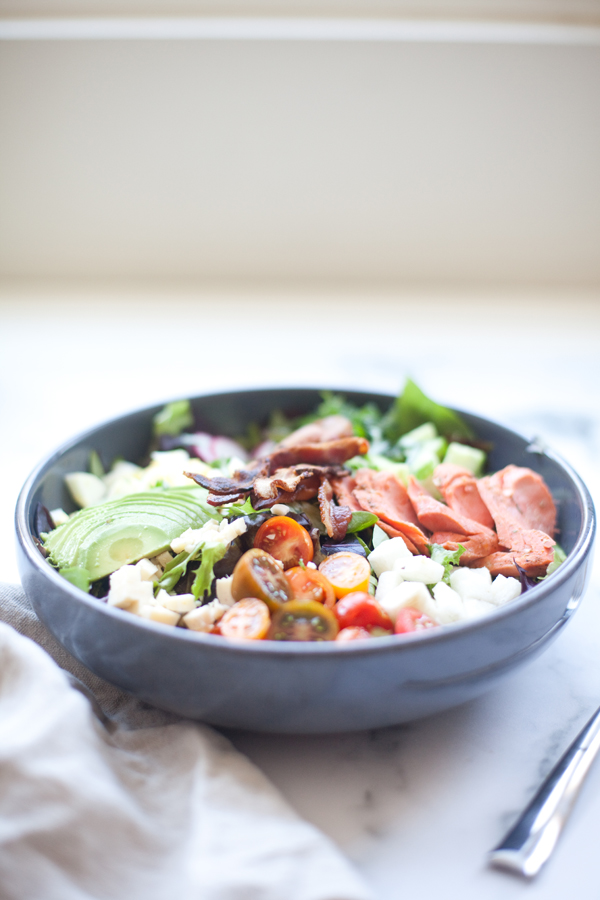 A Blackened Salmon Cobb bowl of salad with sliced avocado, bacon, turkey, cherry tomatoes, cheese cubes, and greens sits on a table next to a fork and napkin.