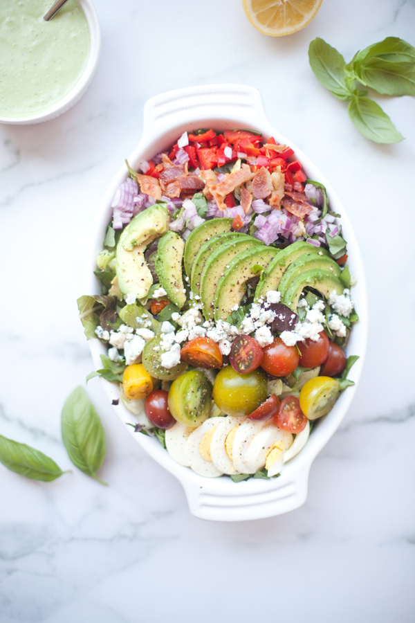 Oval white dish with a colorful Cobb salad featuring avocado, tomatoes, hard-boiled egg, purple onion, red pepper, feta cheese, and greens, garnished with black pepper. The vibrant salad is accompanied by fresh basil leaves, lemon wedges, and a bowl of creamy lemon basil dressing nearby.