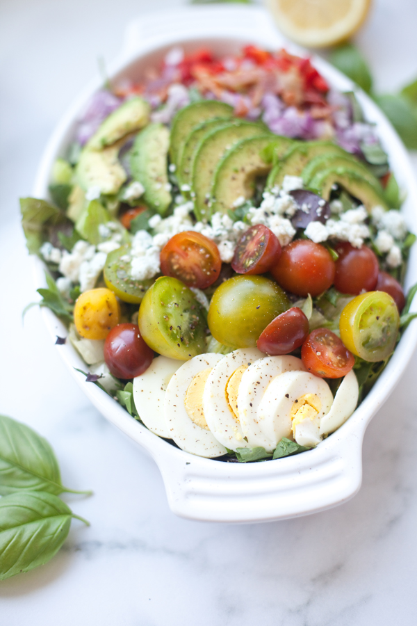 A colorful Cobb salad with creamy lemon basil dressing is served in a white dish, featuring sliced avocado, cherry tomatoes, hard-boiled eggs, crumbled cheese, greens, and chopped red onions. A lemon slice and basil leaves are in the background.