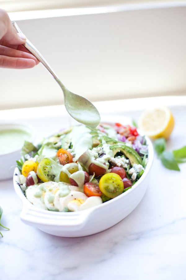 A hand drizzles creamy green dressing over a fresh Cobb salad with cherry tomatoes, avocado, and greens in a white dish. A halved lemon and extra creamy lemon basil dressing sit in the background.