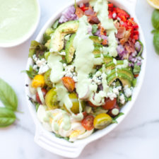 A white dish filled with a colorful Cobb salad with creamy lemon basil dressing, featuring tomatoes, avocado, red onion, feta cheese, and greens—all topped with the vibrant dressing. A bowl of extra creamy lemon basil dressing and fresh basil leaves are nearby.