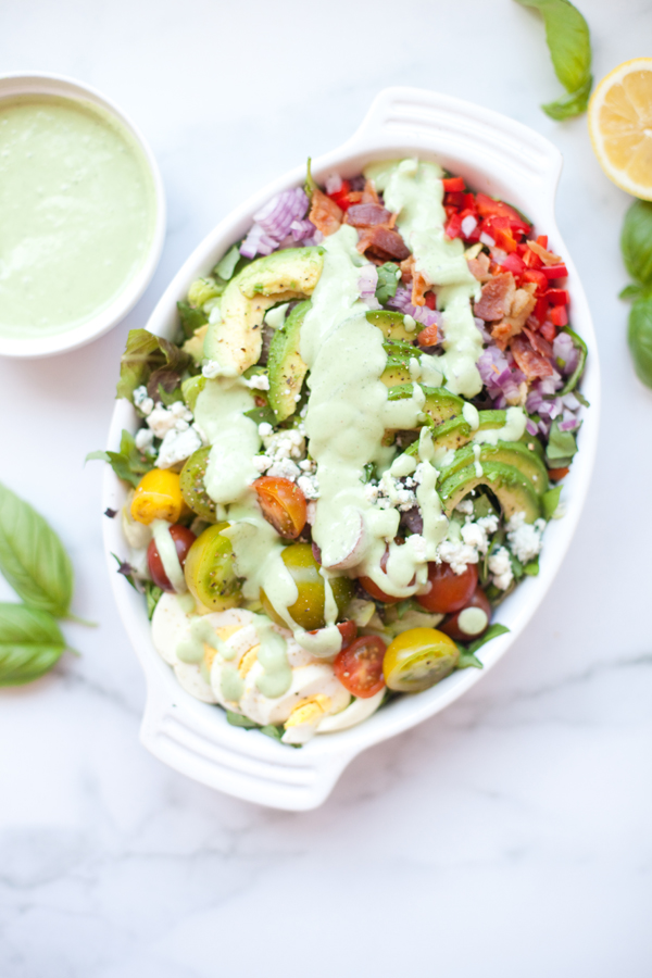 A white dish filled with a colorful Cobb salad with creamy lemon basil dressing, featuring tomatoes, avocado, red onion, feta cheese, and greens—all topped with the vibrant dressing. A bowl of extra creamy lemon basil dressing and fresh basil leaves are nearby.