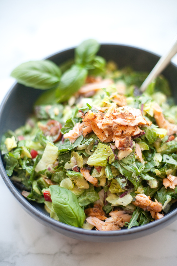 A bowl of chopped salad with greens, shredded salmon, avocado, red onions, and herbs, garnished with fresh basil leaves—reminiscent of a Cobb salad with creamy lemon basil dressing. A fork is resting inside the bowl.