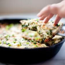A hand lifts a slice of vegetable and turkey mushroom frittata from a black cast iron skillet, with melted cheese and green herbs visible on top.