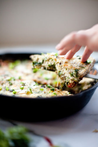 A hand lifts a slice of vegetable and turkey mushroom frittata from a black cast iron skillet, with melted cheese and green herbs visible on top.