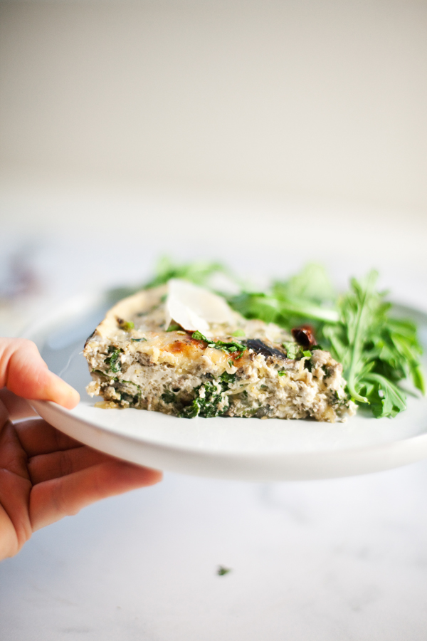 Hand holding a plate with slice of quiche topped with extra arugula.