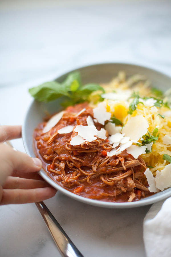 A hand holds a bowl of shredded meat in tomato sauce topped with cheese shavings, served alongside spaghetti squash and fresh herbs—an inviting presentation of Instant Pot Pork Ragu that combines rich flavor with wholesome ingredients.