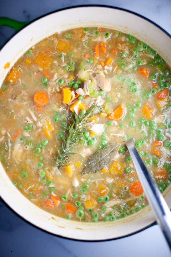 A pot of Veggie Chicken Soup with butternut squash and vegetable medley—carrots, peas, and herbs like rosemary and a bay leaf—simmers invitingly, with a metal spoon inside.