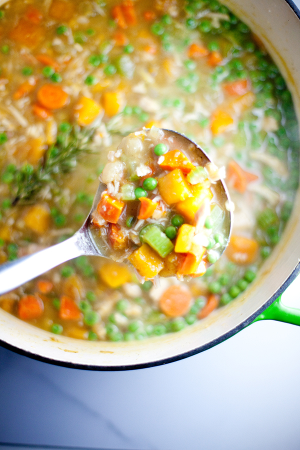 A ladle lifts a serving of hearty Veggie Chicken Soup with butternut squash, chunky carrots, peas, celery, and rice from a large pot, with colorful vegetables and savory broth visible.