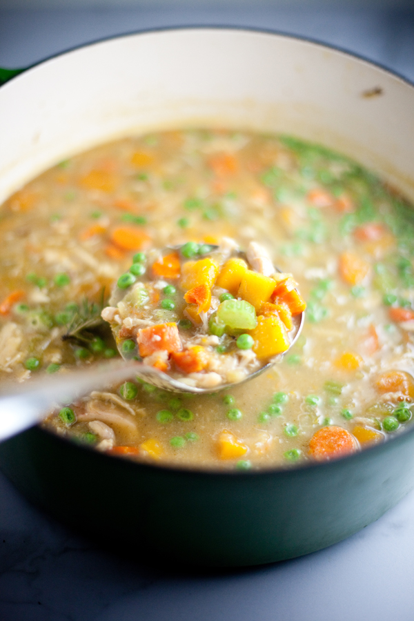 A ladle holds chunky Veggie Chicken Soup with butternut squash, peas, and carrots above a large pot filled with the same hearty soup.