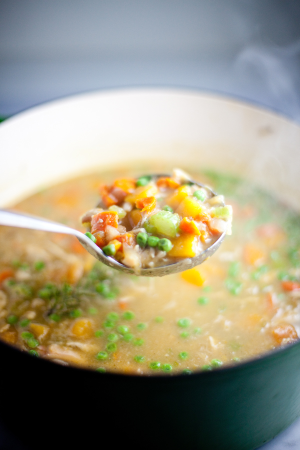 A metal ladle holds a serving of colorful Veggie Chicken Soup with butternut squash above a pot. Steam rises, and the soup contains peas, carrots, celery, and broth.