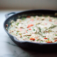 A cast iron skillet filled with Creamy Chicken Pot Pie Soup, featuring visible chunks of chicken, carrots, peas, and garnished with fresh thyme, sits on a light marble surface.