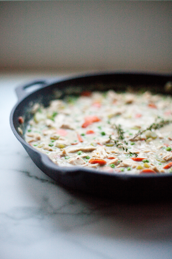 A cast iron skillet filled with Creamy Chicken Pot Pie Soup, featuring visible chunks of chicken, carrots, peas, and garnished with fresh thyme, sits on a light marble surface.