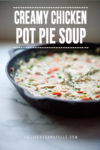 A cast iron pan filled with Creamy Chicken Pot Pie Soup sits on a white surface. Text above reads Creamy Chicken Pot Pie Soup. Veggies and herbs are visible in the rich, comforting soup. Website lillieeatsandtells.com at the bottom.
