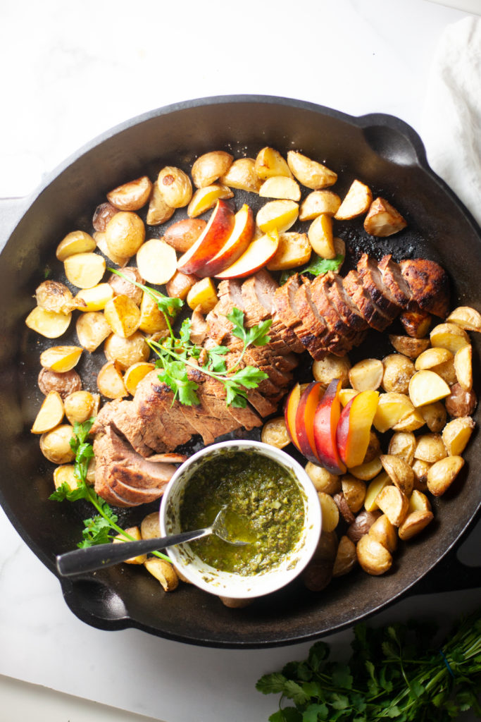 A cast iron skillet with roasted potatoes, sliced pork tenderloin, apple slices, fresh herbs, and a small bowl of green chimichurri sauce with a spoon—the perfect example of Lillie Eats and Tells New Cookbook. Enjoy a healthy twist on single dish comforts in every bite.