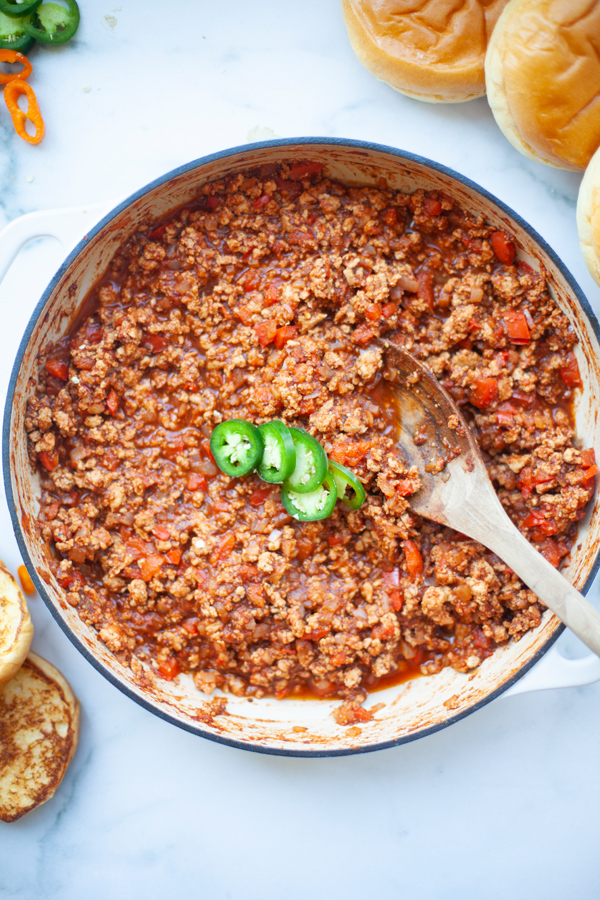 A pot of sloppy joe meat mixture, inspired by Lillie Eats and Tells New Cookbook, is garnished with sliced jalapeños and sits on a white surface, surrounded by a wooden spoon and sandwich buns—a healthy twist on single dish comforts.