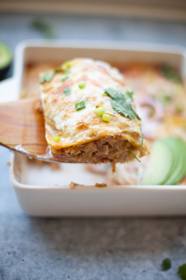 A close-up of a cheesy enchilada being lifted from a white baking dish with a wooden spatula, garnished with green onions and cilantro, with sliced avocado nearby—perfectly capturing the healthy twist on single dish comforts featured in Lillie Eats and Tells New Cookbook.