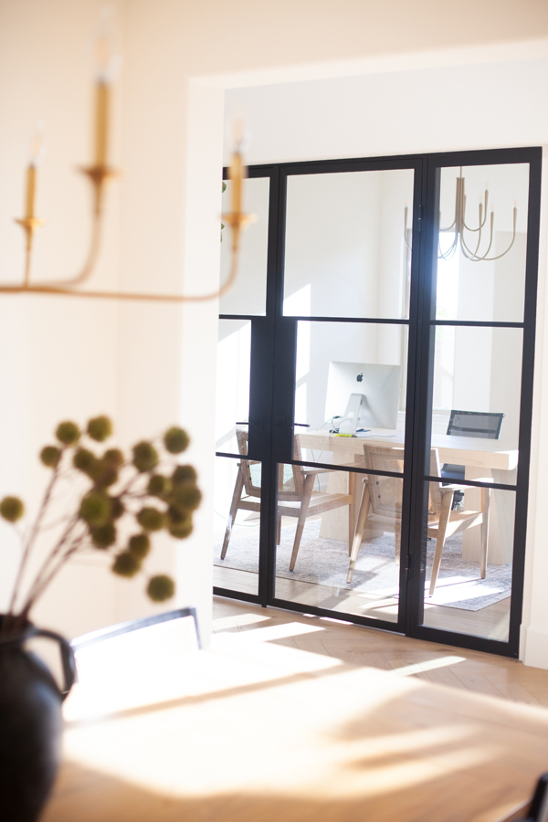Bright home office with glass doors by Pinkys Iron Doors, wooden desk, two chairs, a computer, and a laptop. Modern chandeliers and soft natural light create a warm, inviting atmosphere.