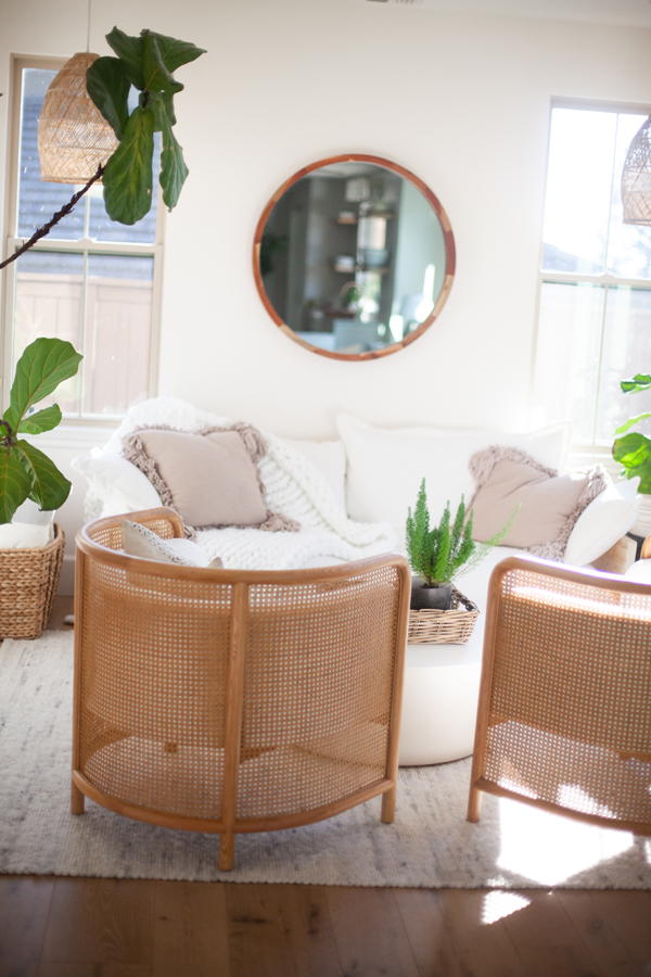 Bright living room with two wicker chairs, a white sofa with neutral cushions and a cozy blanket, a round wall mirror, indoor plants, and natural light streaming through large Pinkys Iron Doors.