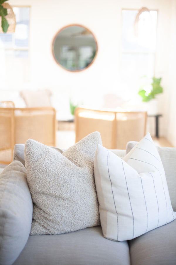 A bright living room with a light gray sofa featuring two textured pillows, cane-back chairs in the background, large windows framed by elegant Pinkys Iron Doors, a round mirror, and a green potted plant.