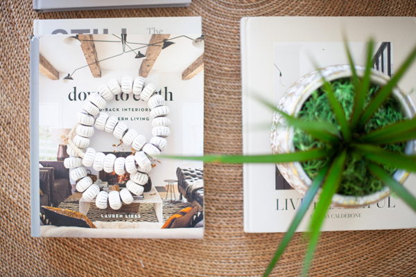 A white beaded necklace rests on a book with an interior design cover, inspired by the elegant aesthetics of Pinkys Iron Doors; beside it is a potted plant and another book, all arranged on a woven surface.