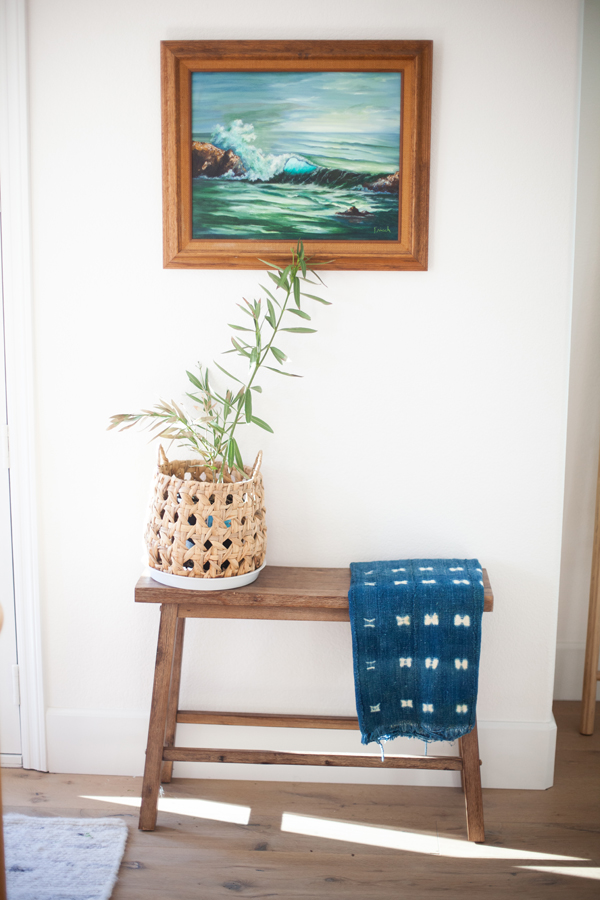 A wooden bench from Pinkys Iron Doors holds a potted plant in a woven basket and a blue patterned cloth. Above, a framed painting of ocean waves hangs on a white wall.