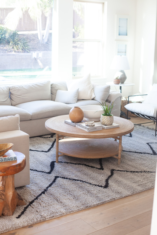 Bright living room with a light gray sectional sofa, round wooden coffee table with decor, modern armchair, and patterned rug. Large windows by Pinkys Iron Doors let in natural light, with a pool visible outside.