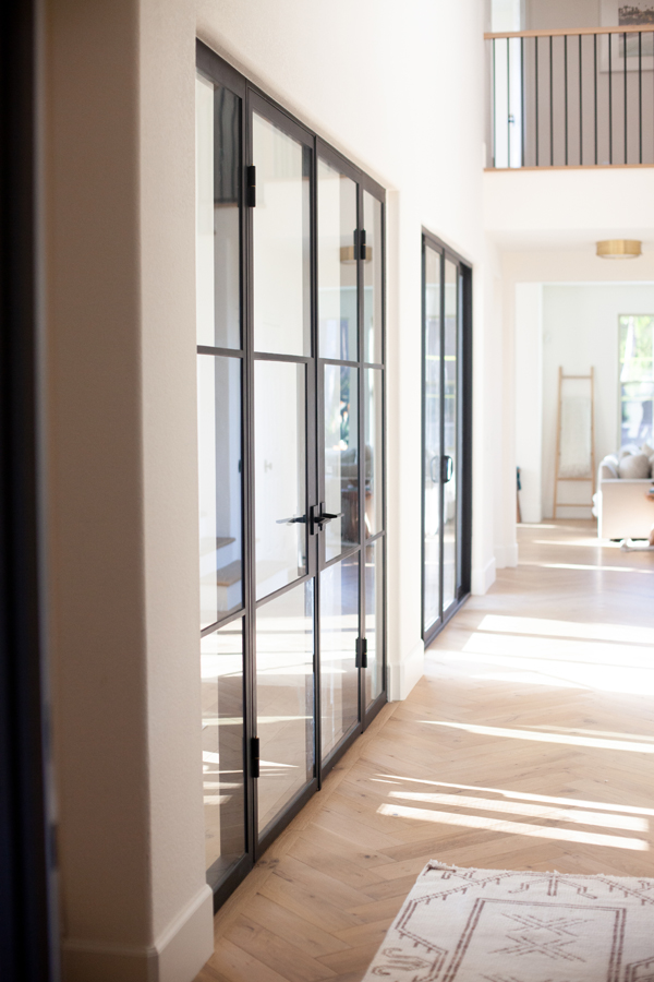 Bright hallway with glass doors by Pinkys Iron Doors, featuring striking black frames and a light wood herringbone floor. Sunlight streams in, casting gentle shadows. A white rug is in the foreground, with a living area and balcony visible in the background.