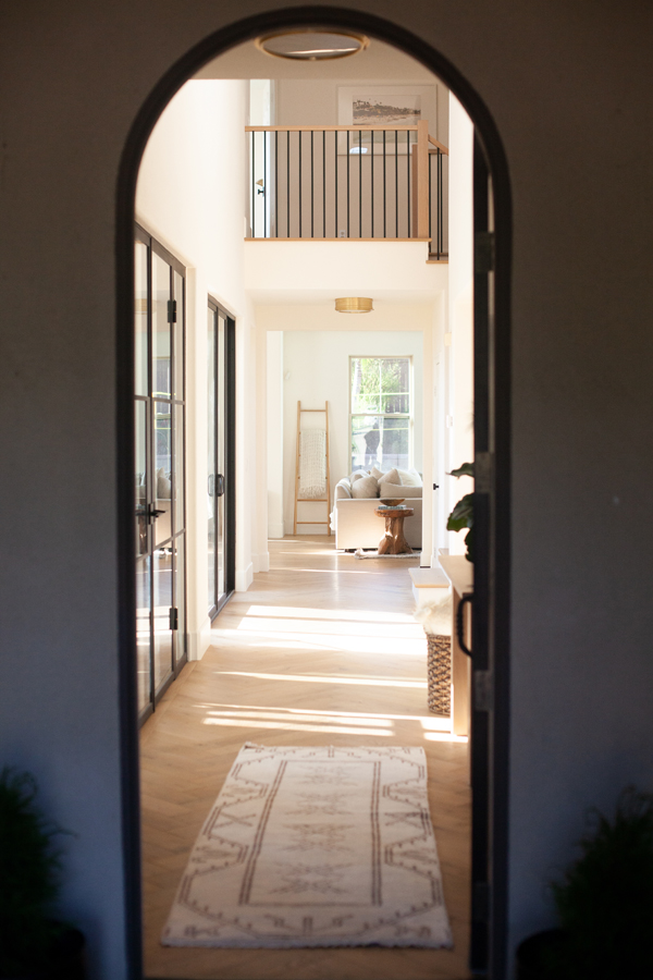 A view through an arched Pinkys Iron Doors doorway into a bright hallway with glass doors, a patterned rug, and a sunlit living area with a sofa, chair, and large mirror in the background.