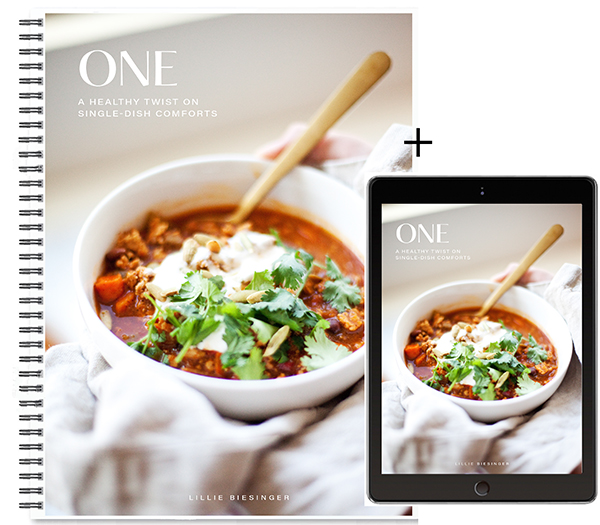 A spiral-bound cookbook and a tablet display the same cover image: a bowl of chili topped with cilantro, sour cream, and nuts. The title reads ONE: A Healthy Twist on Single-Dish Comforts by Lillie Biesinger, featuring delicious recipes from the Lillie Eats and Tells New Cookbook.