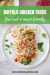 A plate of Buffalo Chicken Tacos topped with slaw, cilantro, and hot sauce sits on a marble surface. A partial drink and extra slaw are visible in the background. Text on the image describes these Buffalo Chicken Tacos as low carb and macro-friendly.
