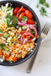 A black bowl filled with shredded chicken, cherry tomatoes, pickled onions, mixed greens, crumbled cheese, and hot sauce—creating an Easy Buffalo Chicken Grain Salad—sits beside a fork and sprigs of cilantro on a marble surface.