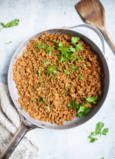 A pan filled with cooked ground turkey seasoned with spices for delicious ground turkey tacos, garnished with fresh cilantro, sits on a light surface with a wooden spatula and a textured kitchen towel nearby.
