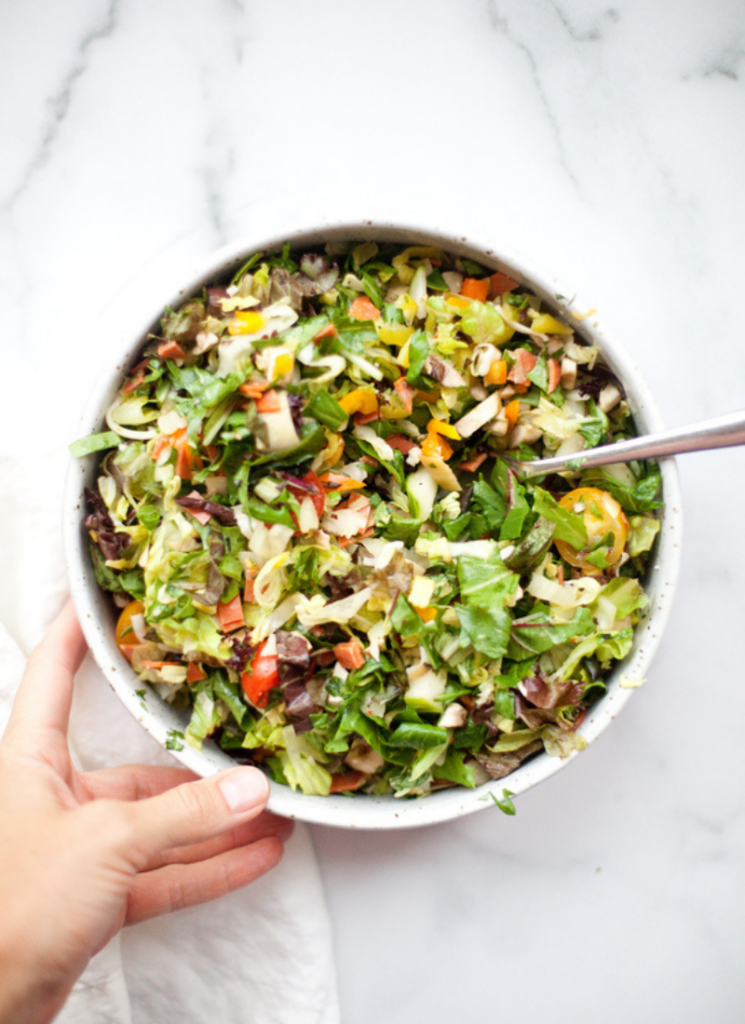A hand holds a bowl of zesty Italian chopped salad with mixed greens, tomatoes, peppers, and onions. A spoon rests inside the bowl on a white marble surface.