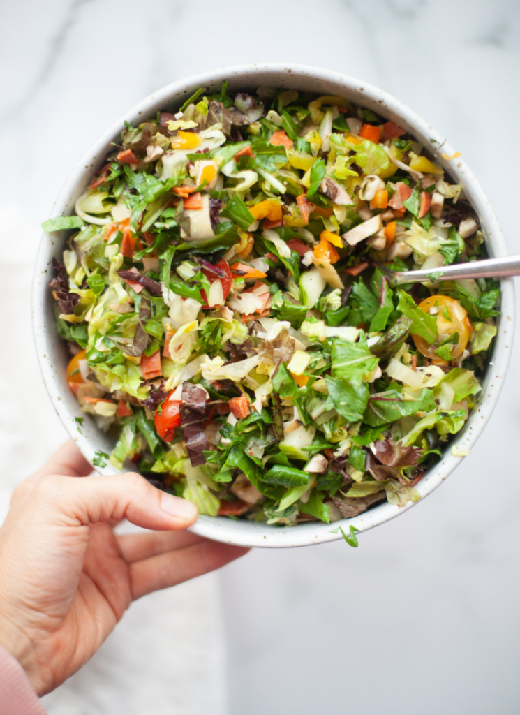 A hand holds a bowl filled with a colorful zesty Italian chopped salad, featuring lettuce, tomatoes, peppers, cucumbers, and other fresh vegetables, with a fork resting inside.