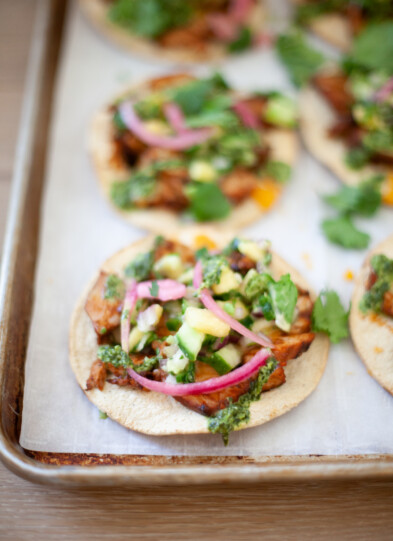 A close-up of Quick BBQ Pork Tostadas topped with shredded meat, chopped vegetables, pickled onions, cilantro, and green sauce on a baking sheet lined with parchment paper.