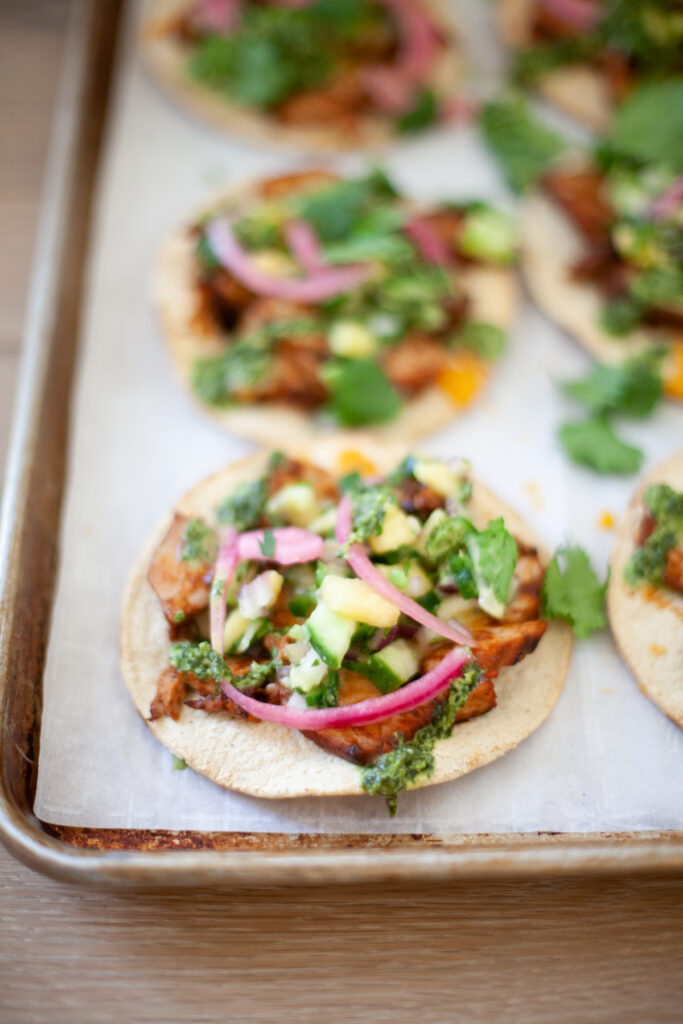 A close-up of Quick BBQ Pork Tostadas topped with shredded meat, chopped vegetables, pickled onions, cilantro, and green sauce on a baking sheet lined with parchment paper.