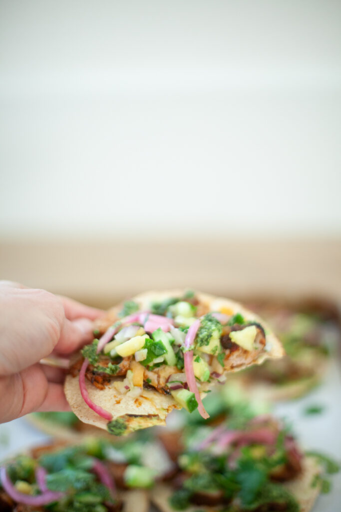 A hand holds a loaded Quick BBQ Pork Tostada topped with chopped vegetables, pickled onions, herbs, and green sauce. More tostadas are blurred in the background on a tray.