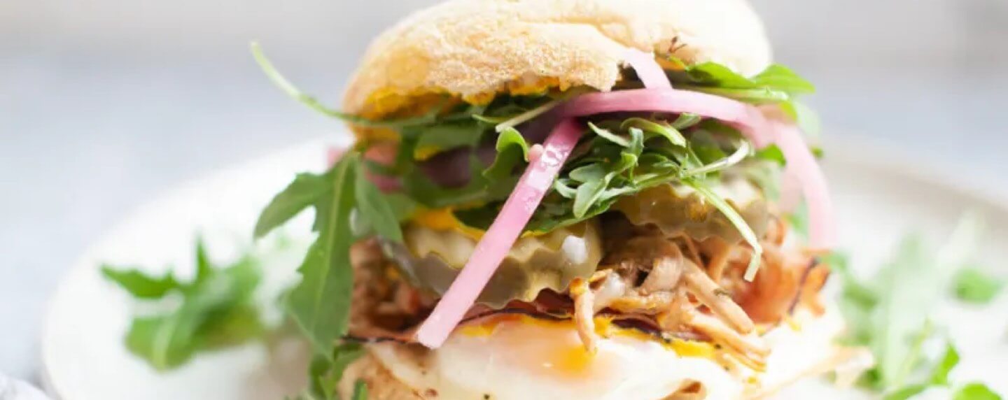 A Cuban breakfast sandwich on an English muffin is filled with pulled meat, melted cheese, pickled onions, pickles, and fresh arugula, served on a white plate.