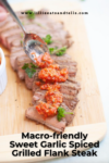 A spoon adds vibrant red garlic sauce to sliced sweet garlic spiced grilled flank steak on a wooden board, garnished with parsley. Text reads Macro-friendly Sweet Garlic Spiced Grilled Flank Steak.